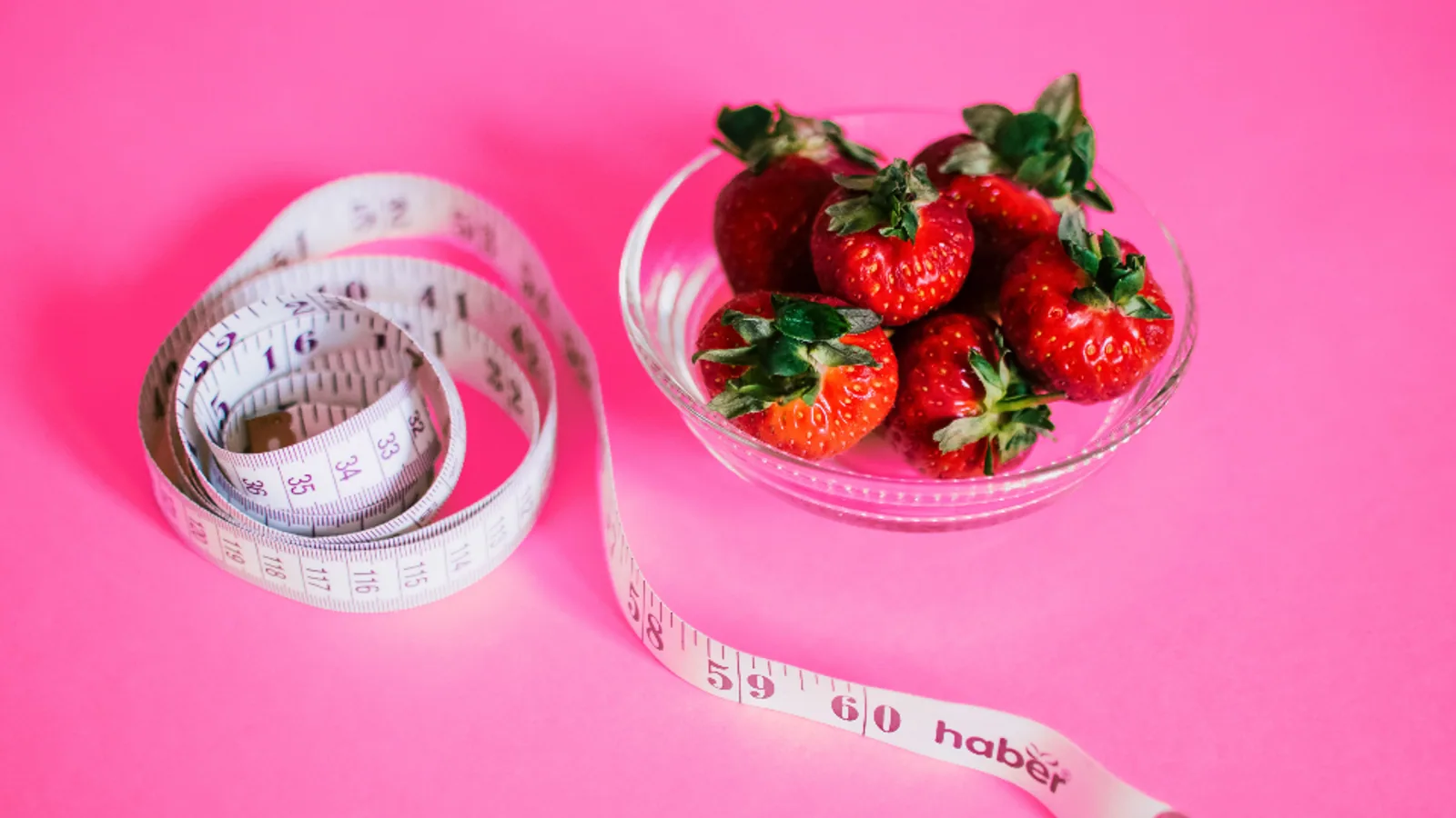Tape measure beside a bowl of strawberries on a pink background