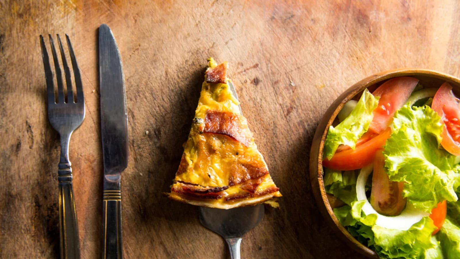 A single pizza slice on a fork beside a bowl of salad